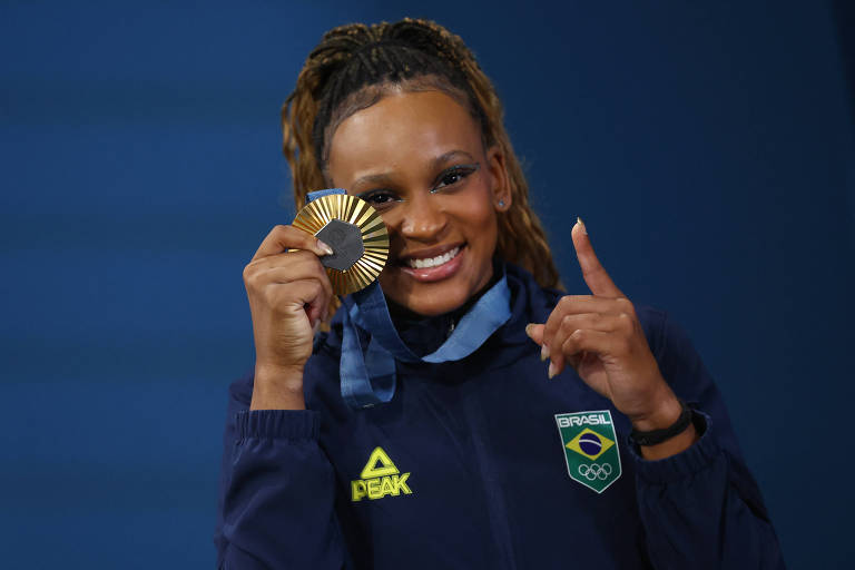 Uma atleta brasileira sorri enquanto segura uma medalha de ouro em uma mão e levanta o dedo indicador da outra mão. Ela está vestindo uma jaqueta azul com o logotipo da marca 'PEAK' e o emblema do Comitê Olímpico Brasileiro. O fundo é azul.