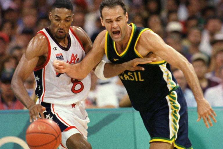 A imagem mostra dois jogadores de basquete em ação durante um jogo. À esquerda, um jogador usa uma camisa branca com detalhes em vermelho e azul, enquanto à direita, um jogador veste um uniforme preto com detalhes em verde e amarelo, representando o Brasil. Ambos estão em movimento, com a bola de basquete visivelmente próxima ao jogador da esquerda, que parece estar tentando driblar. O fundo é desfocado, indicando um ambiente de competição.
