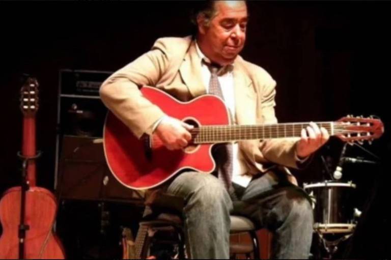 Homem sentado em um palco, tocando um violão acústico vermelho. Ele está vestido com um paletó claro e uma camiseta escura. Ao fundo, há um violão e um tambor. O ambiente parece ser um show ou apresentação musical