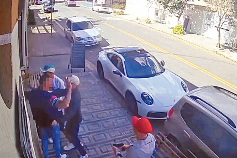 A imagem mostra uma cena de rua capturada por uma câmera de segurança. Três homens, um deles armado, abordam um homem. Ao fundo, há vários carros estacionados, incluindo um carro esportivo branco. 