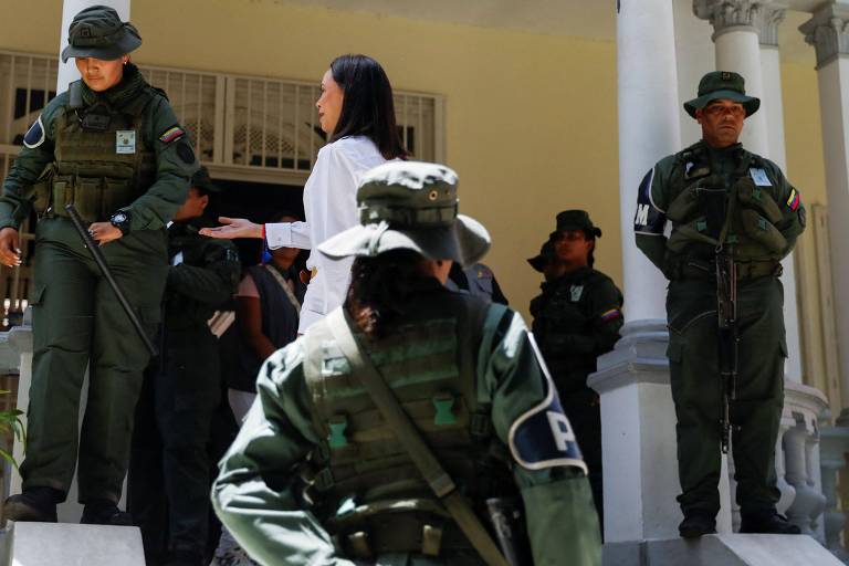 A imagem mostra María Corina estendendo a mão para uma militar que olha para baixo
