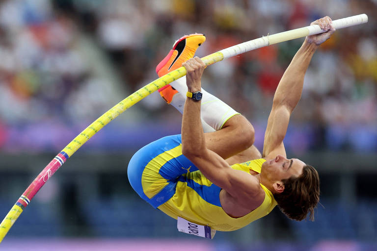 Um atleta está realizando um salto com vara, elevado no ar. Ele usa um uniforme amarelo e azul, e segura a vara com as duas mãos. O fundo mostra uma multidão assistindo ao evento, com várias pessoas visíveis em um estádio.