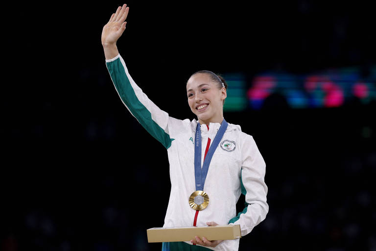 Fotografia tirada de baixo para cima mostra a ginasta franco-argelina Kaylia Nemour no pódio dos Jogos Olímpicos de Paris-2024. Ela usa moletom branco com detalhes verdes, cabelo preso e dá tchau com uma das mãos. No peito, é possível ver a medalha de ouro.