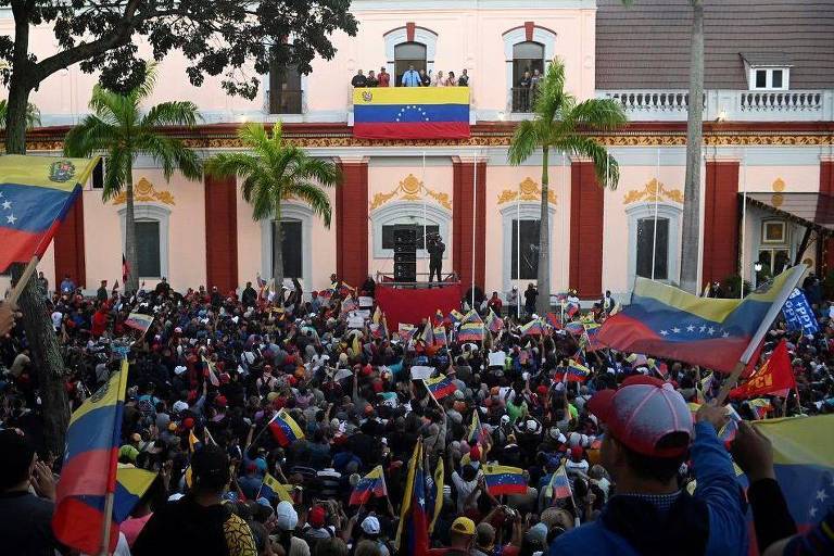 A imagem mostra dezenas de pessoas com bandeiras da Venezuela em frente a um prédio estilo colonial cor de rosa claro. Na sacada central, Maduro fala.