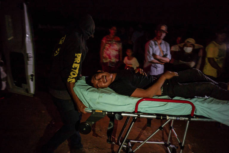 A imagem mostra uma cena de atendimento de emergência em um ambiente com pouca luz. Um homem indígena está deitado em uma maca, chorando de dor, enquanto um socorrista, vestido com uma camisa preta, está ao seu lado. Ao fundo, várias pessoas observam a situação, algumas usando máscaras de proteção. A atmosfera parece tensa