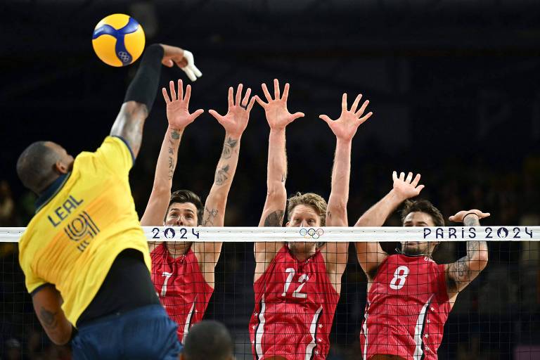 A imagem mostra um jogador de vôlei atacando a bola, enquanto quatro jogadores adversários tentam bloquear o ataque. O jogador que ataca está vestido com uma camisa amarela e preta, e seu nome 'LEAL' está visível nas costas. Os bloqueadores estão em uma rede, levantando as mãos para interceptar a bola. A parte superior da rede exibe as palavras 'PARIS 2024'.
