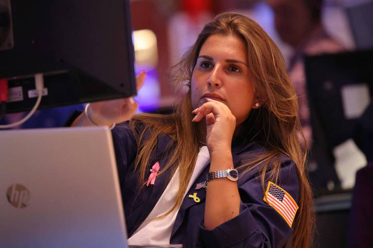 Imagem mostra uma mulher de cabelo longo e castanho claro. Ela usa uma jaqueta azul com o símbolo dos Estados Unidos no ombro. Ela está olhando para duas telas de computador à sua frente com expressão séria.