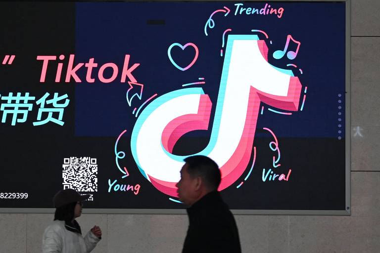 A imagem mostra um grande anúncio digital do TikTok, com o logotipo da plataforma em destaque. O logotipo é estilizado em cores vibrantes, com um fundo escuro. Ao redor do logotipo, há palavras como 'Trending', 'Young' e 'Viral', além de ícones de coração e notas musicais. Na parte inferior, há um código QR e texto em chinês. Uma pessoa caminha em frente ao anúncio.
