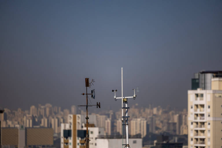 Imagem mostra instrumentos meteorológicos que se parecem com antenas no primeiro plano; há prédios no horizonte e o céu azul está sem nuvens, com uma densa camada cinza na parte baixa, próxima aos prédios 