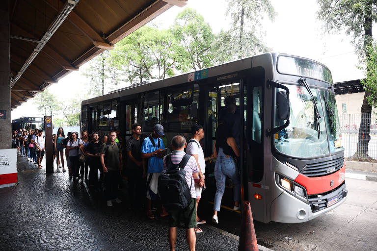 A imagem mostra uma fila de pessoas aguardando para embarcar em um ônibus em uma estação. O ônibus está estacionado próximo à plataforma, e várias pessoas estão na fila, algumas já entrando no veículo. Ao fundo, há árvores e uma estrutura coberta que abriga a estação. O ambiente parece movimentado, com um dia claro.