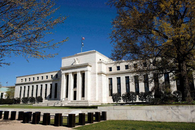 A imagem mostra o edifício da Reserva Federal dos Estados Unidos, com uma fachada de mármore branco e colunas na entrada. O céu está limpo e azul, e há árvores ao redor do edifício. A bandeira dos EUA está hasteada no topo do prédio.
