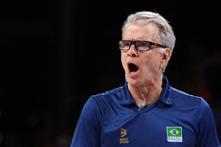 Um homem com cabelo grisalho e óculos, vestindo uma camisa azul com o logo do Brasil, está gritando ou se expressando intensamente durante um jogo de vôlei.
