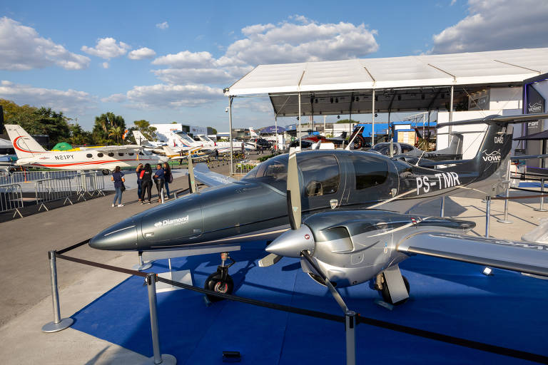 A imagem mostra um avião pequeno de cor cinza escuro em uma exposição ao ar livre. O avião está posicionado em um espaço com um tapete azul e cercado por uma grade. Ao fundo, há outros aviões estacionados e pessoas caminhando. O céu está parcialmente nublado.