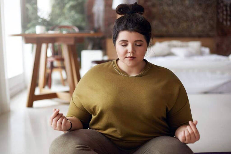 Mulher branca sentada no chão com os olhos fechados. As mãos estão fechadas, apoiadas sobre os joelhos, em posição de meditação.