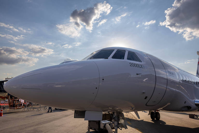 A imagem mostra a parte frontal de um avião, com o céu ao fundo, onde nuvens estão visíveis. A luz do sol está atrás do avião, criando um efeito de silhueta. O avião é de cor branca e está estacionado em uma pista de aeroporto.