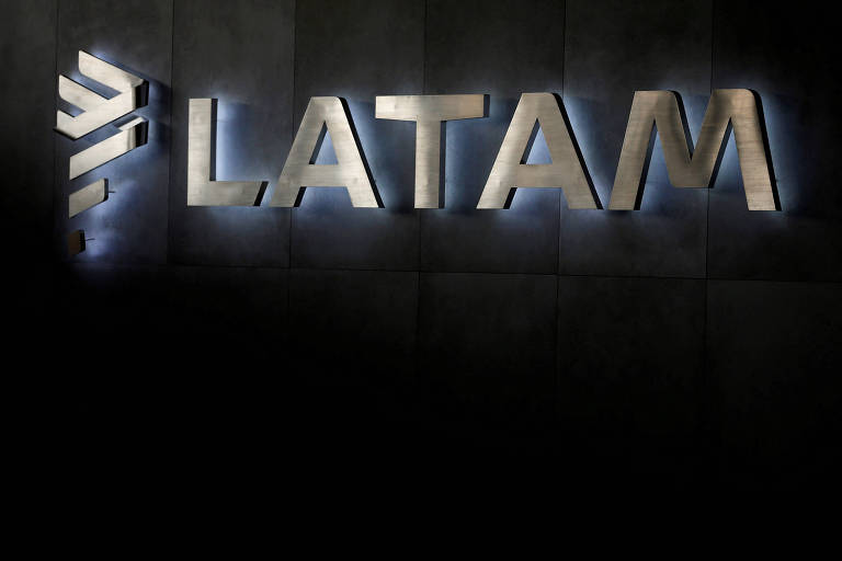 A imagem mostra o logotipo da LATAM, uma companhia aérea, em letras grandes e metálicas. O fundo é escuro, e há uma iluminação suave ao redor das letras, destacando-as. O logotipo é composto pela palavra 'LATAM' em letras maiúsculas, com um símbolo estilizado à esquerda.
