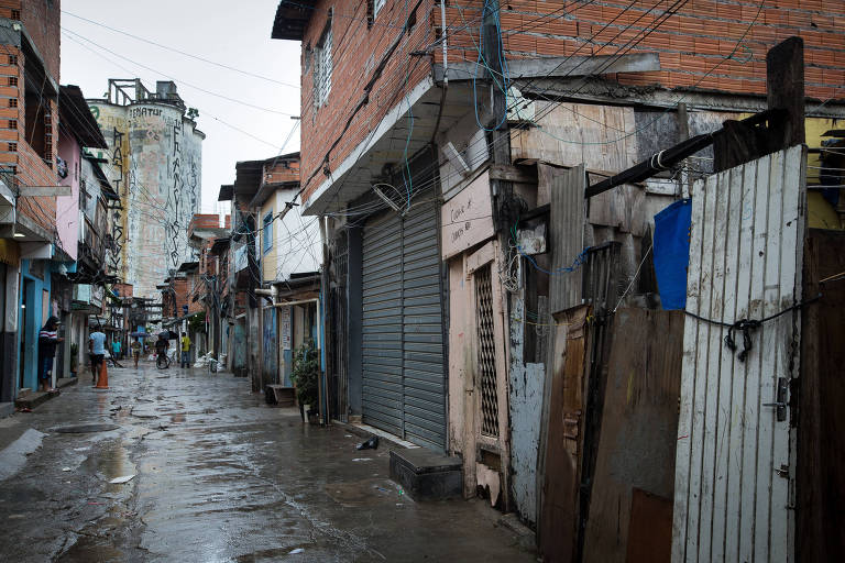 A imagem mostra uma rua estreita em uma área urbana, com casas de tijolos expostos e paredes de madeira. O chão está molhado, indicando que choveu recentemente. À esquerda, algumas pessoas estão visíveis, e ao fundo, um prédio alto com uma pintura visível. Fios elétricos estão pendurados ao longo da rua.