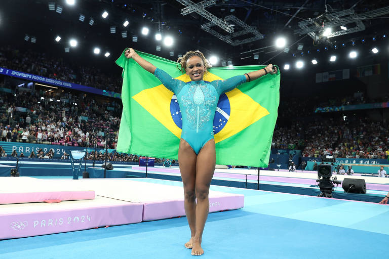 Uma ginasta está em uma competição, segurando a bandeira do Brasil acima da cabeça. Ela usa um traje de ginástica azul e está em uma área de competição com um público ao fundo. O ambiente é iluminado, com luzes brilhantes e equipamentos de ginástica visíveis, como um solo de competição e colchões.