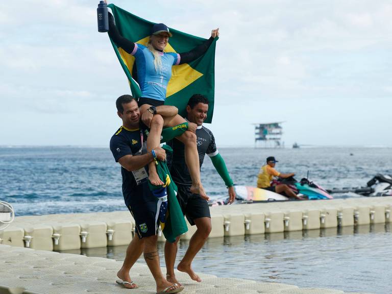 Três pessoas estão em uma área próxima ao mar. Um homem carrega uma mulher nas costas, que segura uma bandeira do Brasil. Outro homem está ao lado, sorrindo. Ao fundo, há uma estrutura flutuante e algumas pessoas em jet skis.
