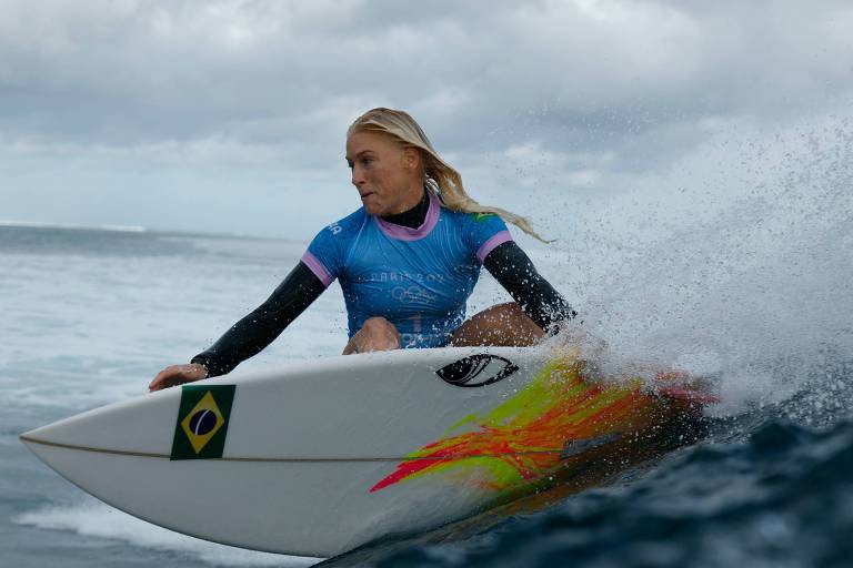 Uma surfista está em cima de uma prancha de surfe, pegando uma onda. Ela usa um traje de neoprene azul com detalhes em rosa e tem cabelo loiro solto. A prancha é branca com uma bandeira do Brasil e um design colorido na lateral. O mar está agitado e o céu apresenta nuvens.
