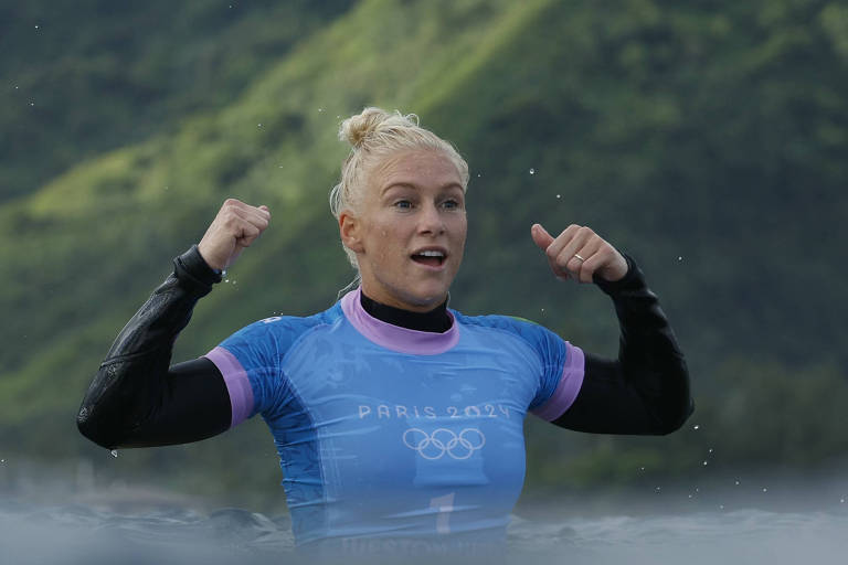 Uma surfista está na água, com um sorriso no rosto e os braços levantados em sinal de celebração. Ela usa uma roupa de neoprene azul com detalhes em rosa e o logotipo dos Jogos Olímpicos de Paris 2024. Ao fundo, há uma paisagem montanhosa verde.
