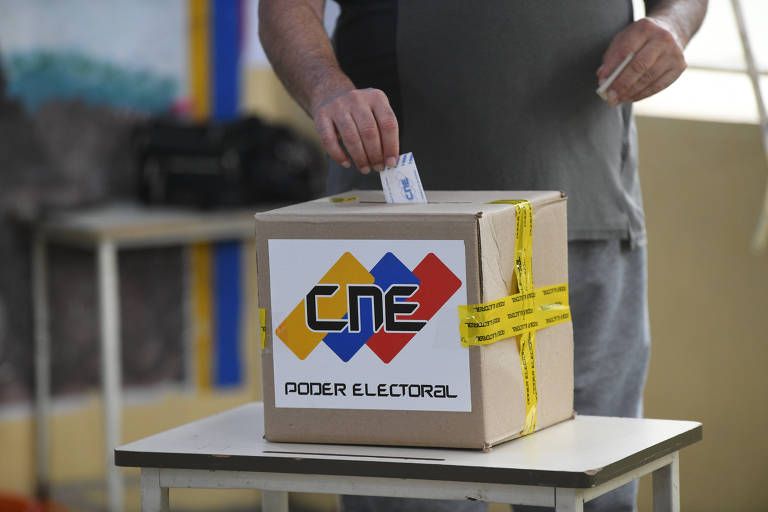 A imagem mostra uma urna eleitoral em uma mesa. Uma pessoa está inserindo um voto na urna, que é de cor marrom e possui o logotipo do CNE (Conselho Nacional Eleitoral) em destaque. A urna está amarrada com uma fita amarela e há um fundo desfocado que sugere um ambiente de votação.