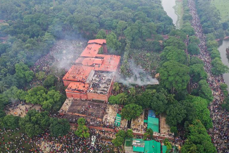 A imagem mostra uma vista aérea de um incêndio em um edifício de tijolos vermelhos cercado por vegetação densa. Há fumaça saindo do telhado do edifício, e uma grande multidão de pessoas se reúne nas proximidades. À direita, há uma área com árvores e um rio visível. O cenário é de um evento significativo, com muitas pessoas visíveis ao longo de um caminho.