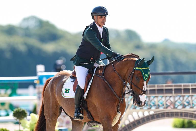 Um cavaleiro montado em um cavalo, ambos em um ambiente de competição equestre. O cavaleiro usa um colete de competição preto e calças brancas, além de um capacete. O cavalo é de pelagem castanha e está equipado com uma cabeçada verde. Ao fundo, há uma pista de salto e uma vegetação ao longe.