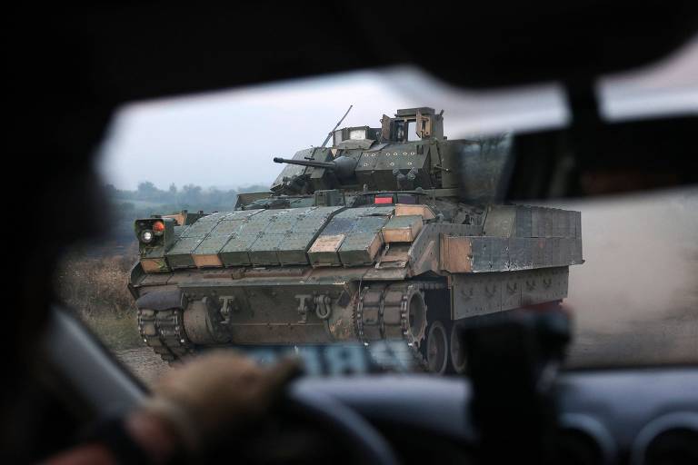 Uma veículo militar blindado, camuflado de verde e marrom, com um canhão, é visto de dentro de um carro civil numa estrada