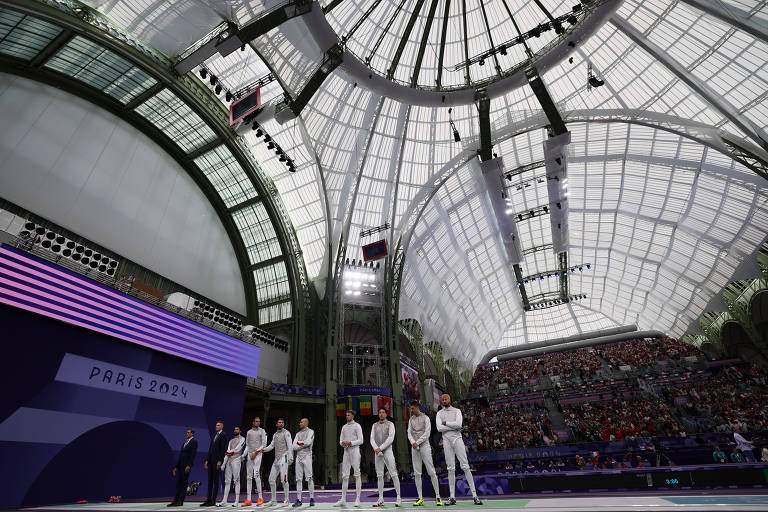 A imagem mostra um grupo de atletas em trajes esportivos alinhados em um palco, sob uma grande estrutura de vidro e metal. O ambiente é iluminado e há uma multidão visível ao fundo, com arquibancadas cheias. O teto é alto e apresenta um design arquitetônico moderno.