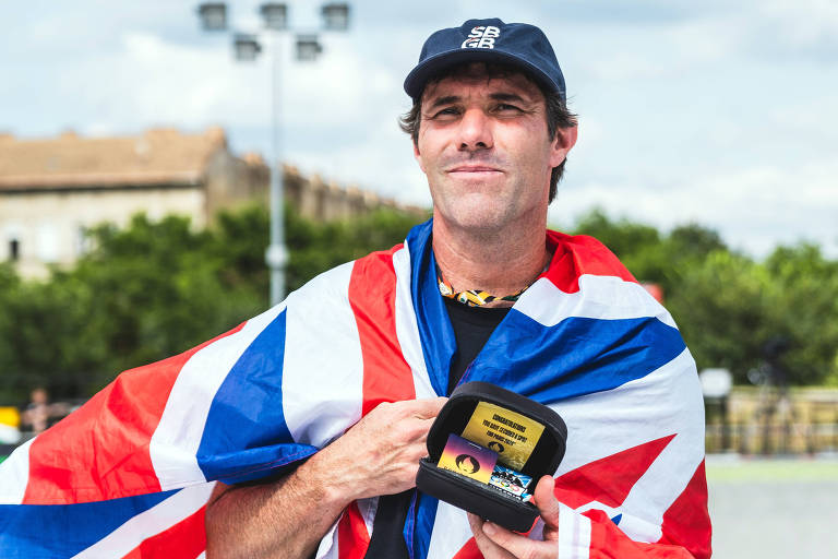 Um skatista está posando com um sorriso, envolto em uma bandeira britânica. Ele usa um boné azul escuro e segura uma caixa preta com um prêmio dentro. Ao fundo, há um parque de skate e um céu parcialmente nublado.
