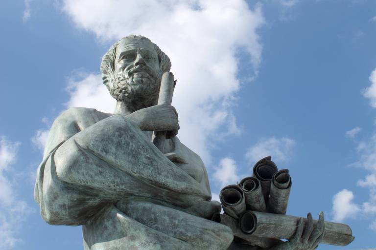 A imagem mostra uma estátua de um filósofo com barba, segurando um rolo de pergaminho em uma das mãos e vários outros rolos embaixo do braço. O fundo é composto por um céu azul com algumas nuvens brancas.
