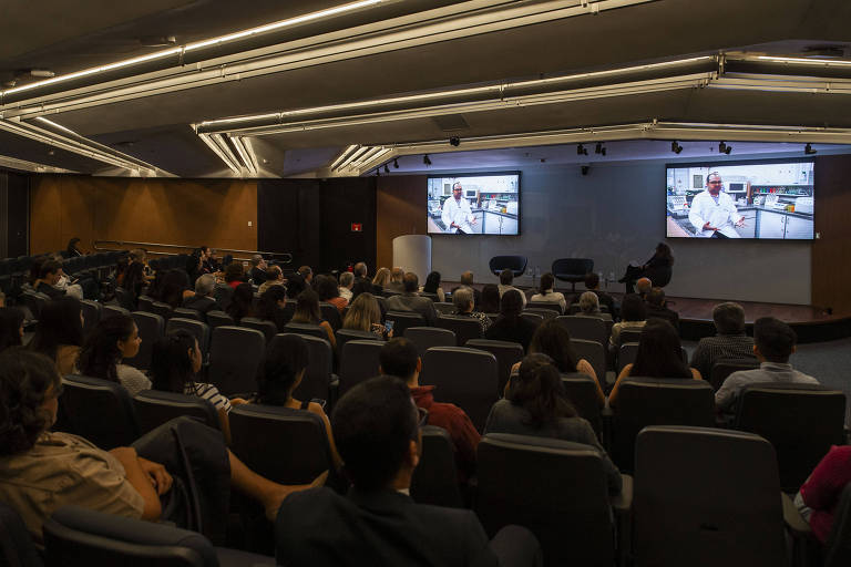 Auditório cheio com pessoas sentadas em cadeiras. Em frente a ele, um palco, em que estão sentadas três pessoas. Há dois televisores grandes nas pontas do palco.