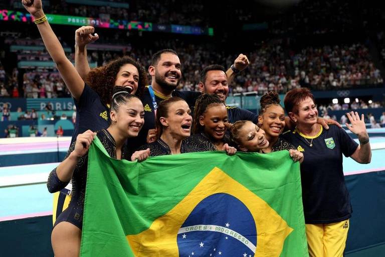 Um grupo de atletas e membros da equipe de ginástica brasileira posam juntos, sorrindo e segurando a bandeira do Brasil. Eles estão em um ambiente de competição, com um público visível ao fundo. A equipe parece estar celebrando uma conquista, com expressões de alegria e entusiasmo.