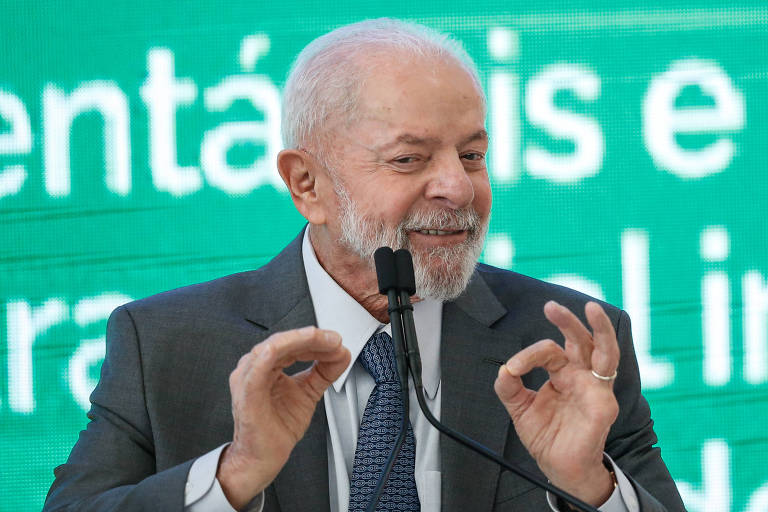 Lula sorri e gesticula, de terno e gravata diante de um microfone. ele tem barba e cabelos ralos grisalhos. o fundo é verde