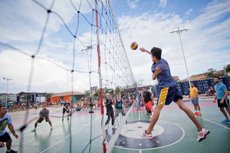 A imagem mostra um jogo de vôlei em uma quadra ao ar livre. Um jogador está saltando para atacar com a bola em direção ao lado adversário, enquanto outros jogadores estão em movimento ao redor. A quadra é cercada por uma rede e há várias pessoas jogando. O céu está parcialmente nublado.