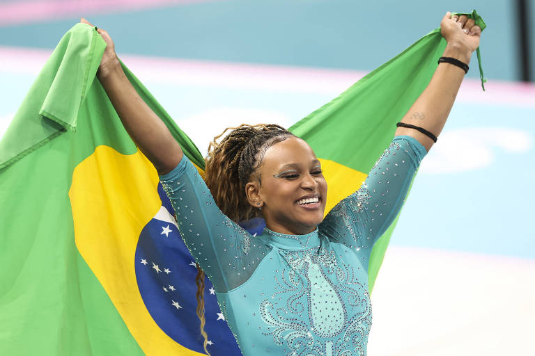 Sorrindo e segurando a bandeira do Brasil às costas， com as duas mãos， Rebeca Andrade comemora a medalha de ouro no solo da ginástica artística nos Jogos Olímpicos de Paris