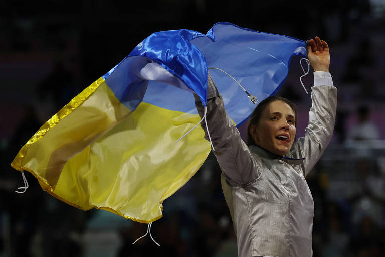 Uma atleta está celebrando com uma bandeira da Ucrânia, que é azul e amarela. Ela está vestindo um traje prateado e levantou a bandeira acima da cabeça, sorrindo. O fundo mostra uma multidão desfocada, sugerindo um evento esportivo.