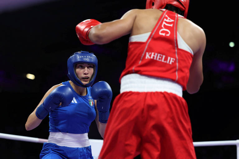 A imagem mostra duas boxeadoras em um ringue. A atleta à esquerda está vestindo um uniforme azul com um capacete de proteção azul, enquanto a atleta à direita está usando um uniforme vermelho com o nome 'KHELIF' visível. Ambas estão em posição de combate, com a boxeadora de azul parecendo estar em uma postura defensiva, enquanto a de vermelho está avançando.
