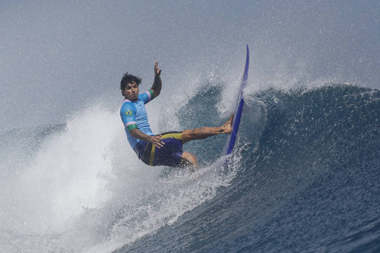 Um surfista está realizando uma manobra em uma onda. Ele usa uma camisa azul e shorts escuros, enquanto se equilibra em uma prancha de surf azul. A água está espumando ao redor dele, e o céu parece nublado ao fundo.