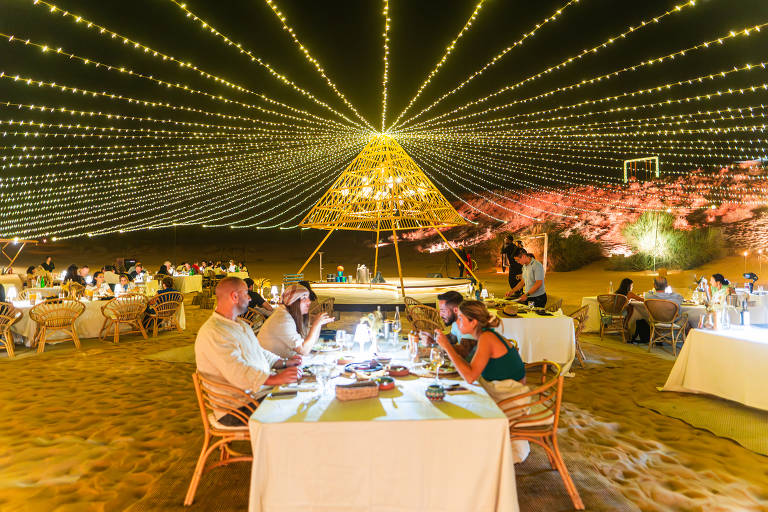 A imagem mostra um jantar ao ar livre em um ambiente desértico à noite, com mesas dispostas na areia. As mesas estão decoradas com toalhas brancas e há pessoas sentadas, algumas conversando e outras comendo. Acima, há uma iluminação decorativa com luzes penduradas em cordas, criando um efeito de estrela. No fundo, uma estrutura em forma de pirâmide feita de bambu é iluminada, e há vegetação ao redor.