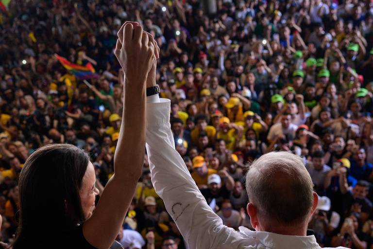 A imagem mostra duas pessoas de costas, levantando as mãos unidas em um evento público. Ao fundo, uma multidão animada, com pessoas segurando bandeiras e usando chapéus amarelos e verdes. A atmosfera é de celebração e apoio.