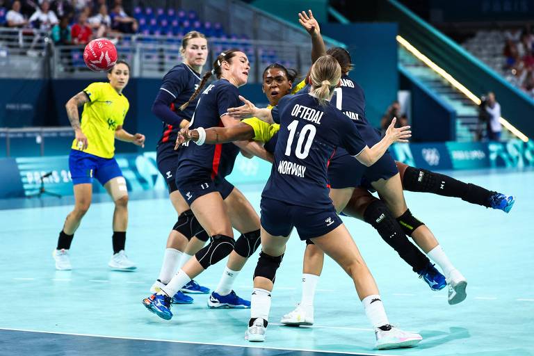 A imagem mostra uma partida de handebol feminino, com jogadoras em ação. As atletas estão vestindo uniformes de cores diferentes, com algumas em azul escuro e uma em amarelo. A jogadora com a camisa número 10 está em destaque, enquanto outras jogadoras estão se movimentando ao redor dela. O ambiente é um ginásio esportivo, com arquibancadas ao fundo.
