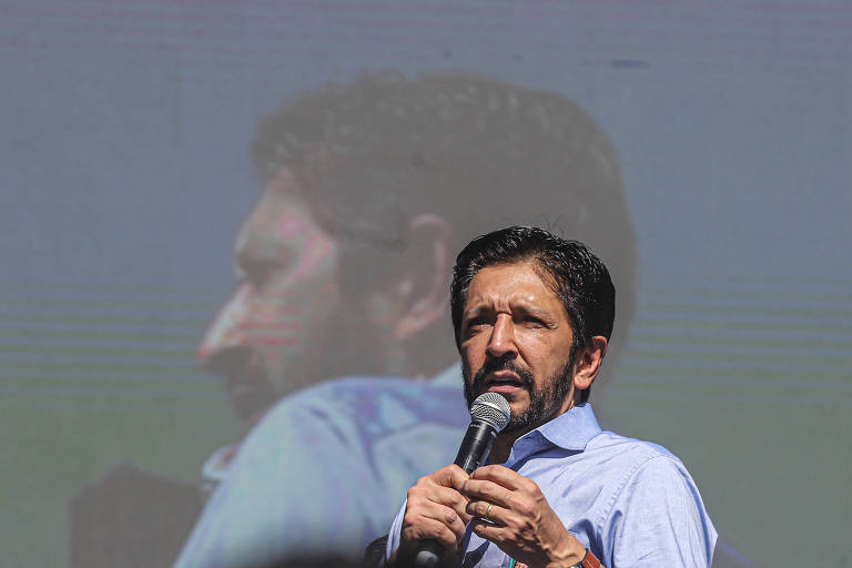 Um homem está falando ao microfone em um evento. Ele usa uma camisa azul e tem cabelo escuro e barba. Ao fundo, há uma projeção de sua imagem em um telão, mostrando seu perfil. O ambiente parece ser ao ar livre, com um fundo desfocado.