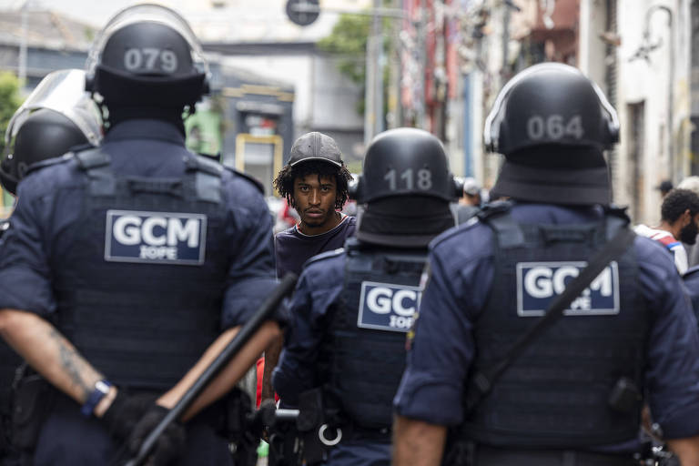 A imagem mostra um grupo de policiais em uniformes táticos, com capacetes e coletes, em uma rua urbana. Eles estão de costas, voltados para um indivíduo à frente. Os policiais têm em seus uniformes a sigla 'GCM'. O ambiente é urbano, com prédios e uma atmosfera de vigilância.
