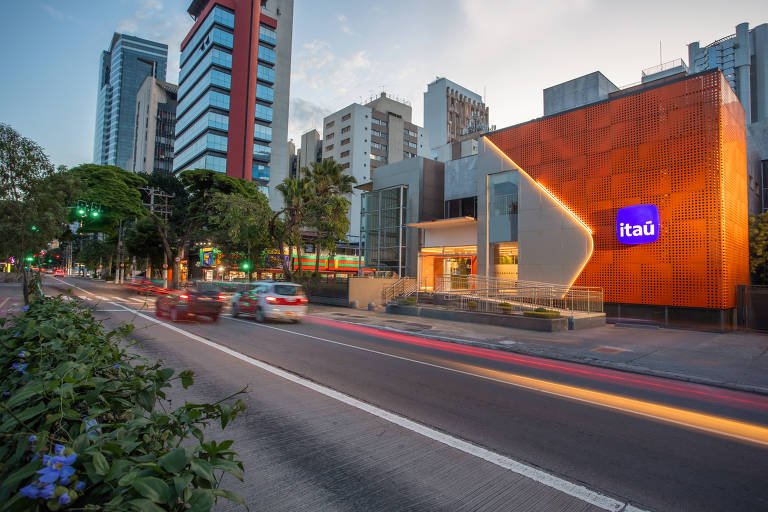 Fachada de agência do Itaú iluminada, com carros e ciclistas passando à frente