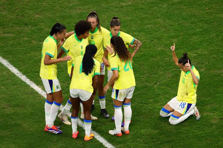 Um grupo de jogadoras de futebol está reunido em campo, vestindo uniformes amarelos. Algumas estão em pé, enquanto uma jogadora está ajoelhada, levantando uma mão. O campo é verde e parece ser um momento de estratégia ou celebração.
