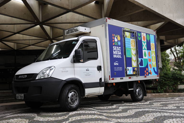 Um caminhão branco da marca Iveco, modelo Daily, estacionado sob uma estrutura de concreto. O veículo possui um compartimento refrigerado na parte traseira, decorado com um design colorido que inclui formas geométricas e elementos gráficos. O lado do caminhão exibe a inscrição 'SESC MESA BRASIL' e informações de contato, como um número de telefone e um site. O chão é coberto por pedras portuguesas dispostas em um padrão ondulado.