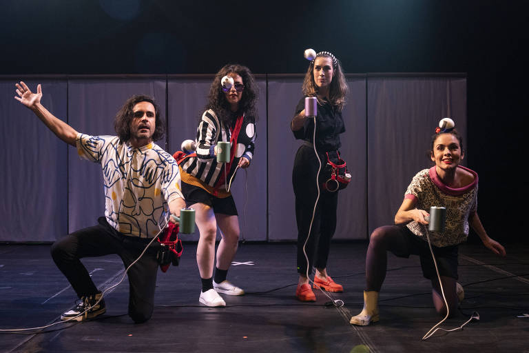 Quatro performers estão em uma cena teatral. O homem à esquerda usa uma camisa estampada e está com o braço levantado. A mulher ao centro, com óculos e uma blusa listrada, segura um copo e parece estar em movimento. A mulher à direita, vestindo uma blusa preta, também segura um copo e está em uma posição de ataque. A última mulher, agachada, usa uma blusa com estampas e sorri enquanto segura um copo. Todos têm objetos brancos em suas cabeças e estão em um palco com fundo escuro.