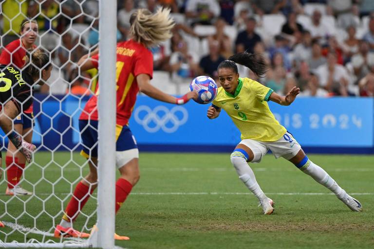 A imagem mostra uma jogadora de futebol vestindo um uniforme amarelo, se preparando para chutar a bola em direção ao gol. Ela está em um campo de futebol, próximo à rede do gol, enquanto uma jogadora adversária, vestindo um uniforme vermelho e azul, tenta bloquear a jogada. O fundo é composto por uma multidão assistindo ao jogo.